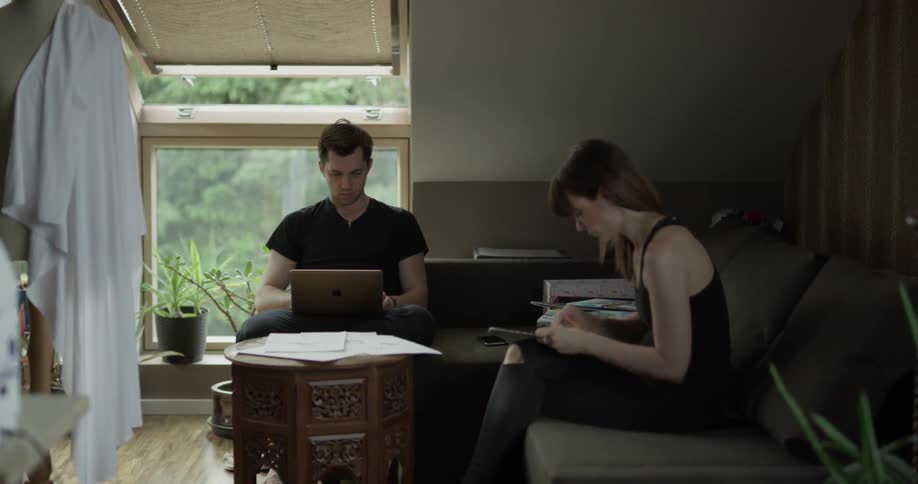 A woman draws on a drawing pad while a man sitting on the same couch types on a laptop