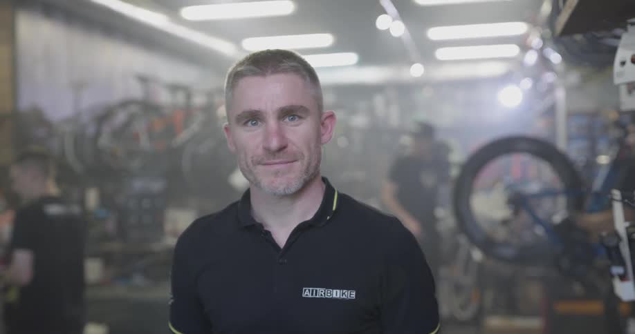 White man smiles at a bicycle repair shop garage or store while a tire spins in the background