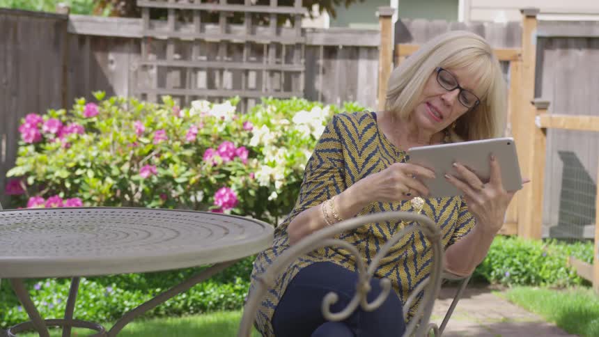 Older woman looks at tablet sitting at table in fenced yard