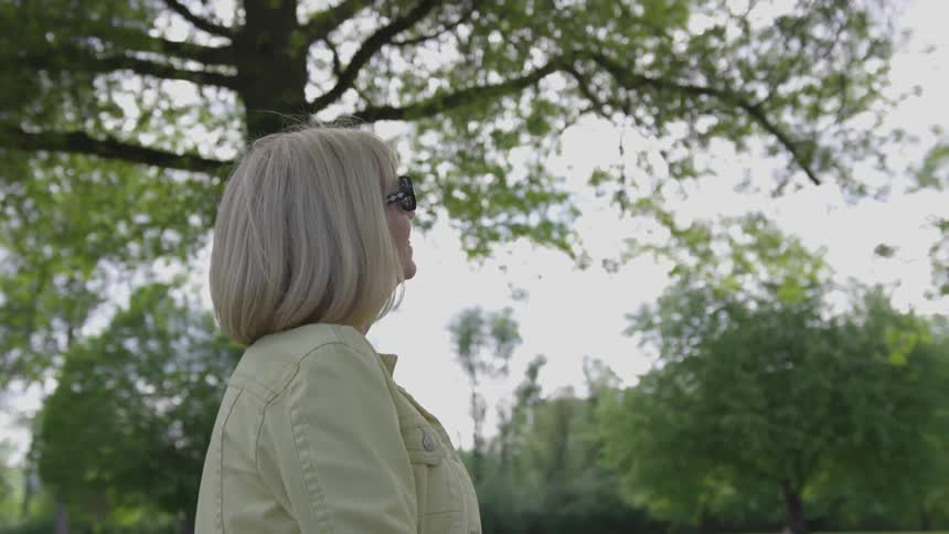 An older woman wears sunglasses as she looks at the sky under a tree