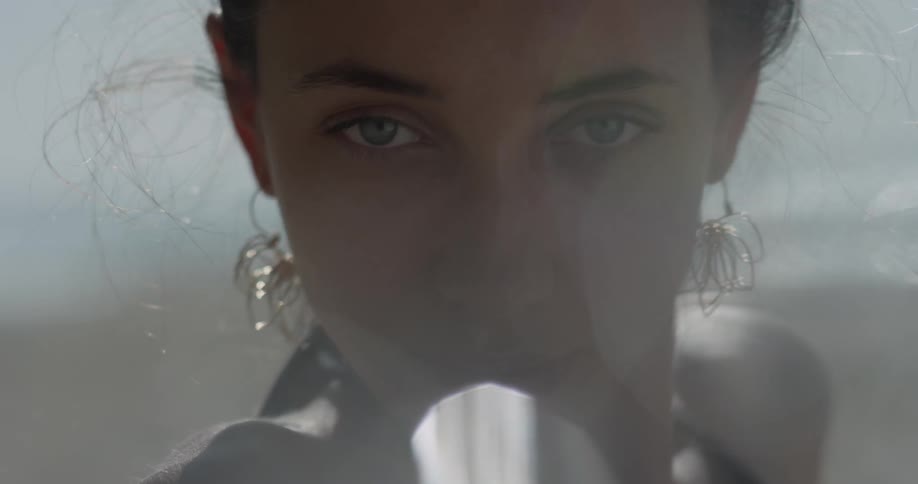 Woman with gold earrings looking into camera and swaying head back and forth while blinking with ocean in background