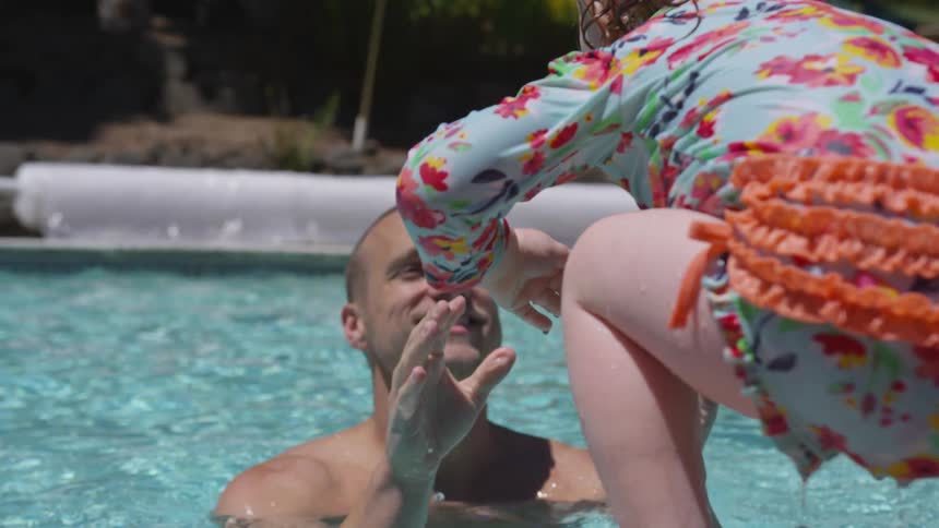 A white male stands in a pool with arms outstretched to catch a female toddler as she jumps in the water