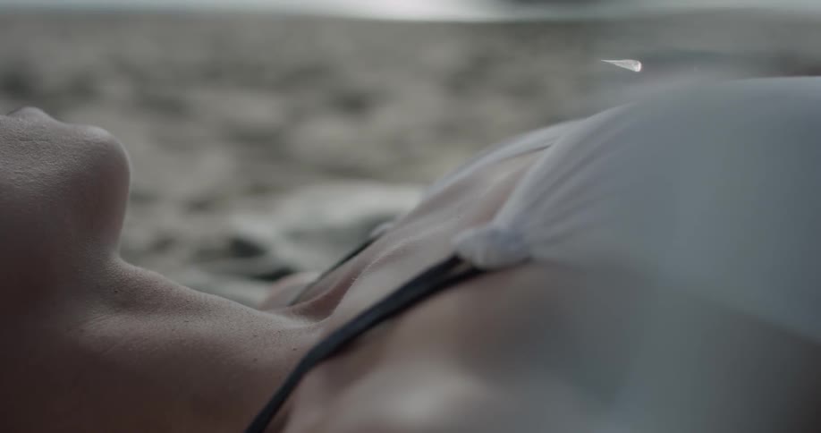 A woman lays in the sand at the beach as waves rush in in the background wearing a white dress with the sunlight on her skin