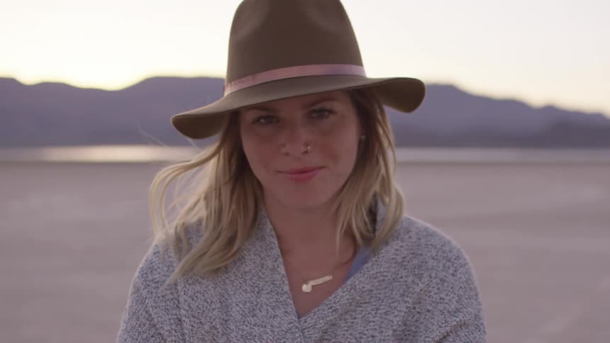 A woman wearing a hat and sweater looks up and smiles at sunset