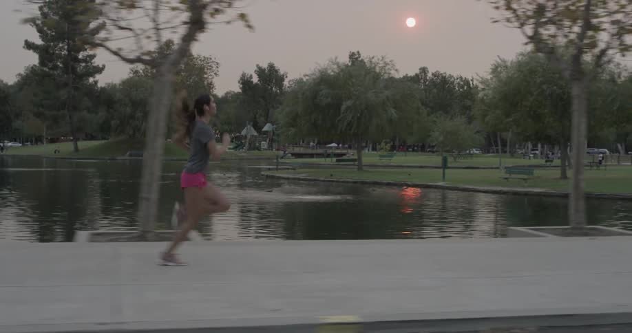 Women in short red shorts runs on the sidewalk at the park with smoke covered skies making the sun visible