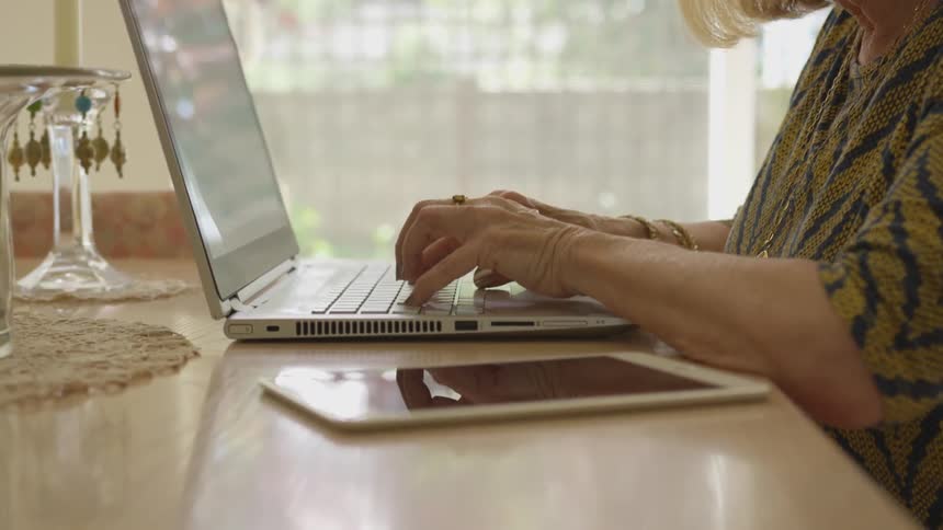 An older woman sits down and types on a laptop indoors at day