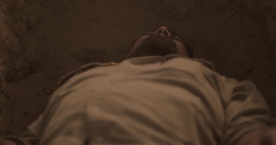 A man lies down in a dirt hole and pulls a gun out of his pocket