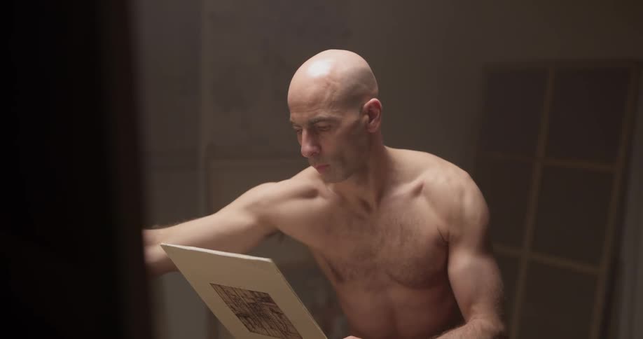 A bald man without a shirt looks at something in his hand then paints on a large canvas