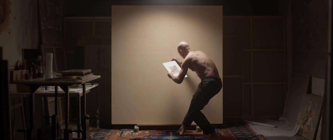 A man draws on a large canvas while looking at a smaller drawing in his other hand
