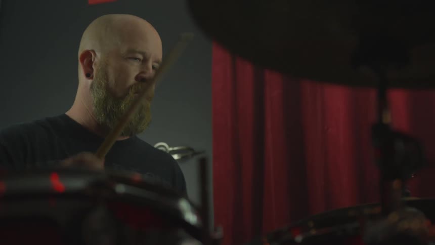 A man plays a drum kit that's sitting in front of red theater curtains