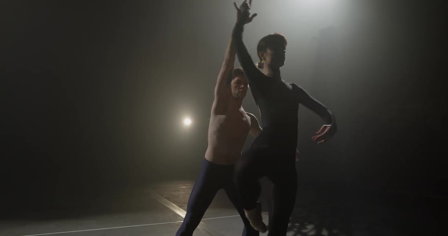 A man and a woman dance on a dark stage illuminated by stage lights
