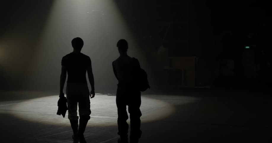 Two men in a dark room with a spotlight walking together holding clothing