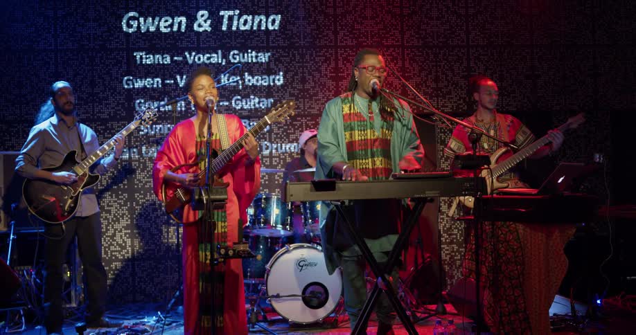 African people play music up on stage in a concert room with red and blue lights and many musical instruments