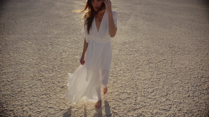 A woman wearing a white dress walking on dry dirt in the sun