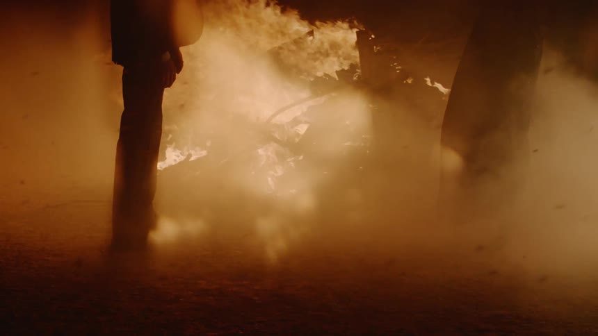 A man and a woman stand on opposite sides of a burning fire
