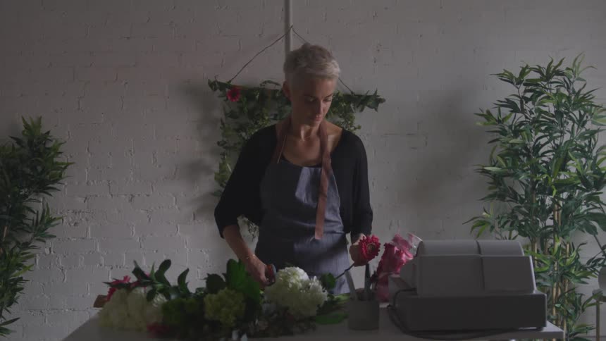 Woman cuts flowers at a table with register in front of brick wall