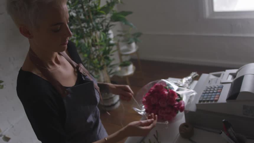 A blonde, short-haired florist arranges a bouquet of flowers next to her till