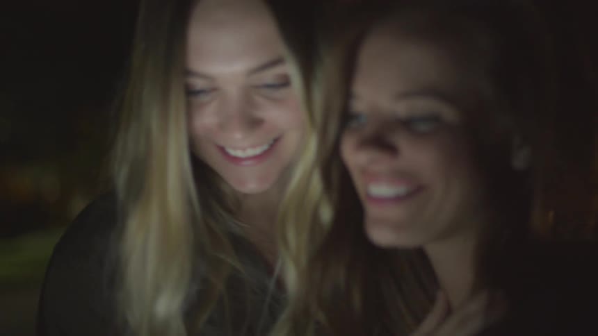 Two women talk and look at their phone at night