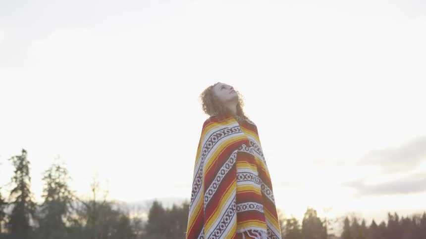 A woman wrapped in a blanket stands outside staring up at the sky