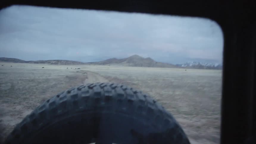 A vehicle with a spare tire drives along a road surrounded by fields