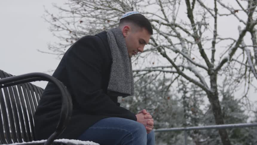 A man wearing a yarmulke, heavy coat and scarf sits outside on a bench on a snowy day