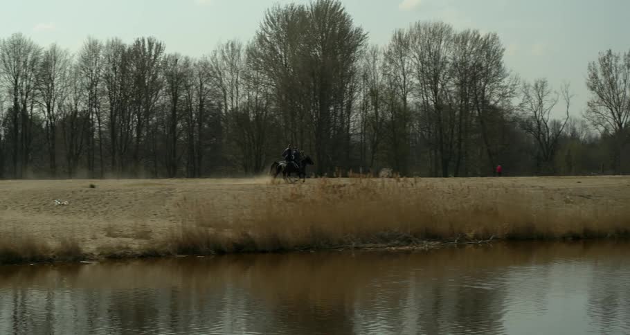 Two people are riding their horses along a river in daylight