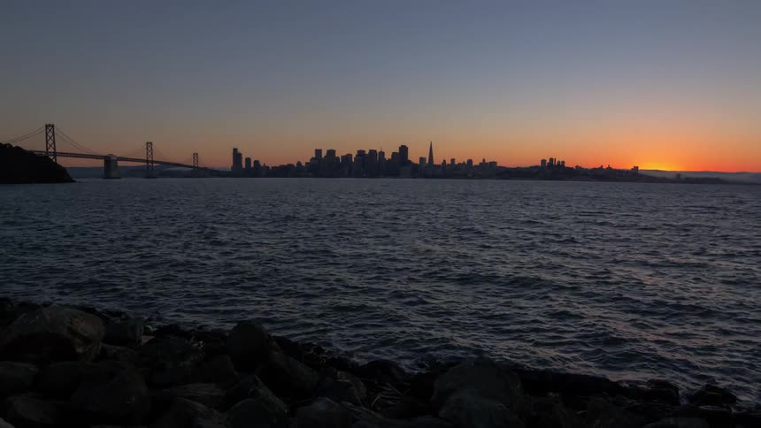 The sun sets and night falls over a city skyline and the ocean