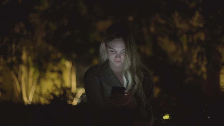 Young woman looks at cell phone and smiles outdoors at night