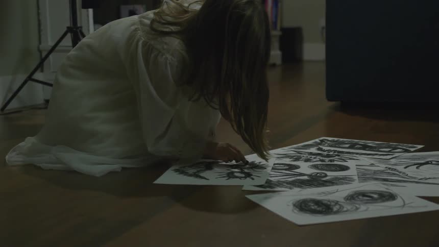 A woman wearing thin white dress is kneeling on the ground of the living room floor drawing psychotic scary sketches on white sheets of paper