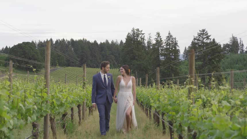 A man in a tuxedo walks with a woman in a dress through a garden