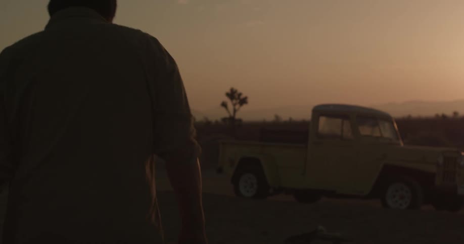 A man walking towards an old truck in the desert at sunset