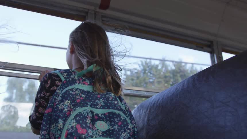 Backside of a young girls upper body as she looks out the wind of the school bus on a sunny day