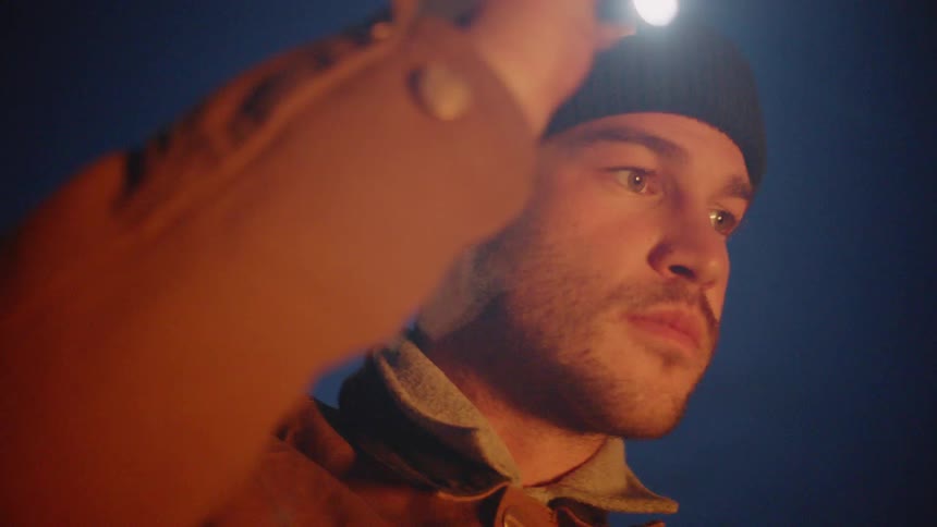 A man in a coat and winter hat holds a shining flashlight in the dark
