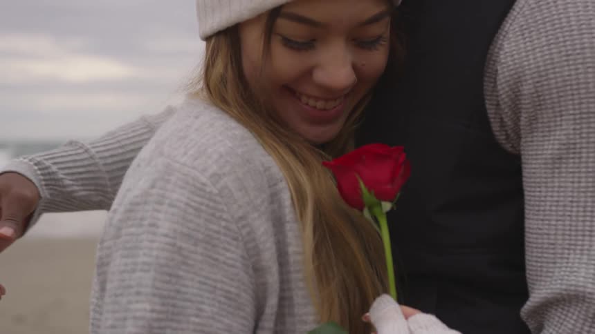 A woman is hugged on a beach while holding a red rose