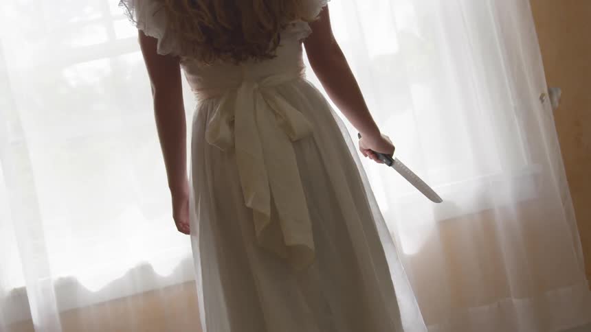 A woman stares out the window with a knife in her hand