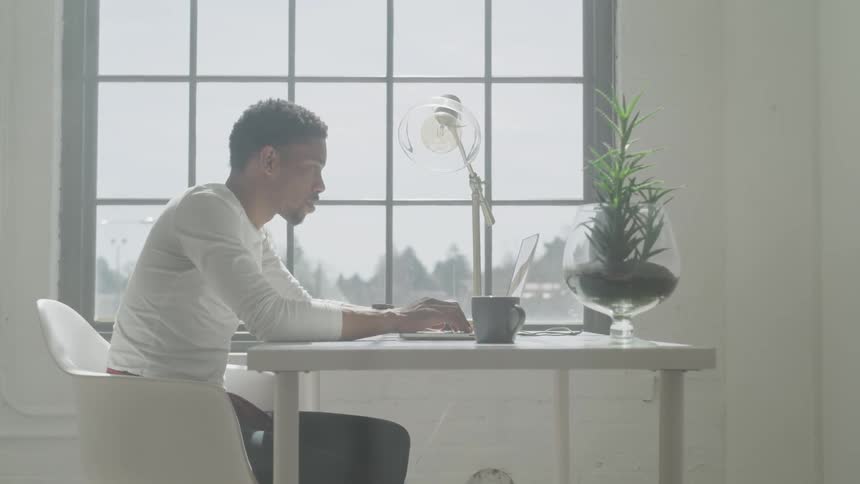 A man works on a laptop at a desk in front of a window
