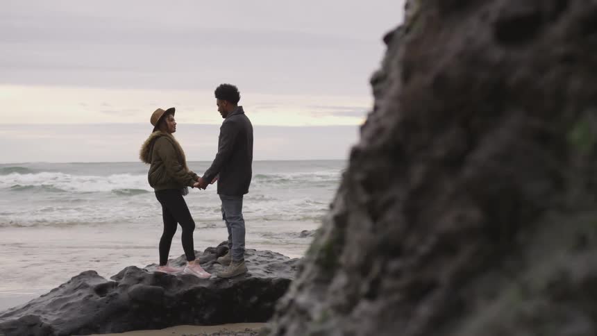 A man and woman hold hands standing on a large rock by the water