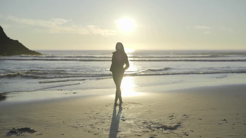Women stands on the beach in front of the water as waves come in as the sun is on the horizon