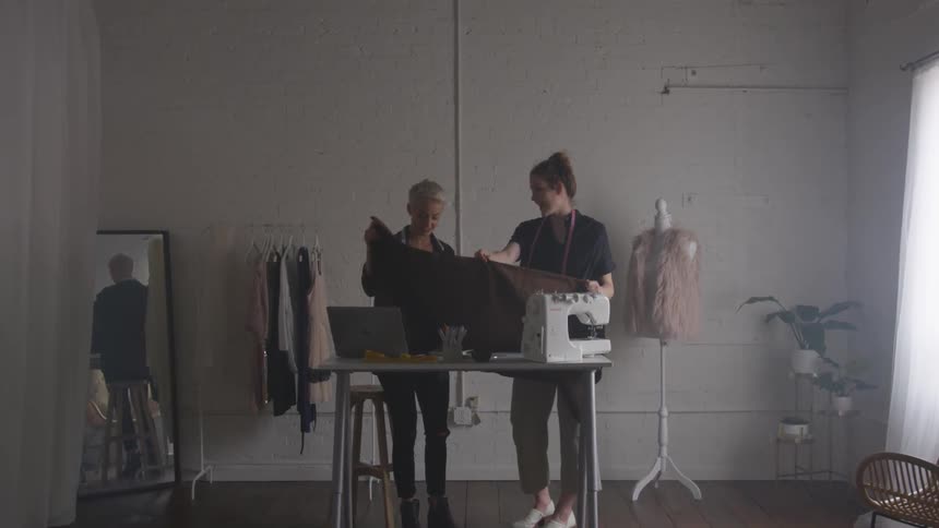 Two women standing in front of a table with a sewing machine as they hold fabric and talk about it