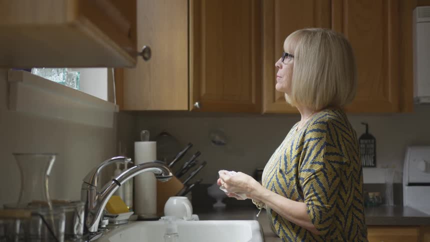 An old woman stands at a sink looking out a window
