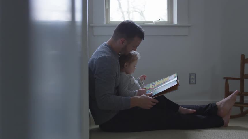 A man sits against a wall beneath a window reading a book with his daughter in his lap