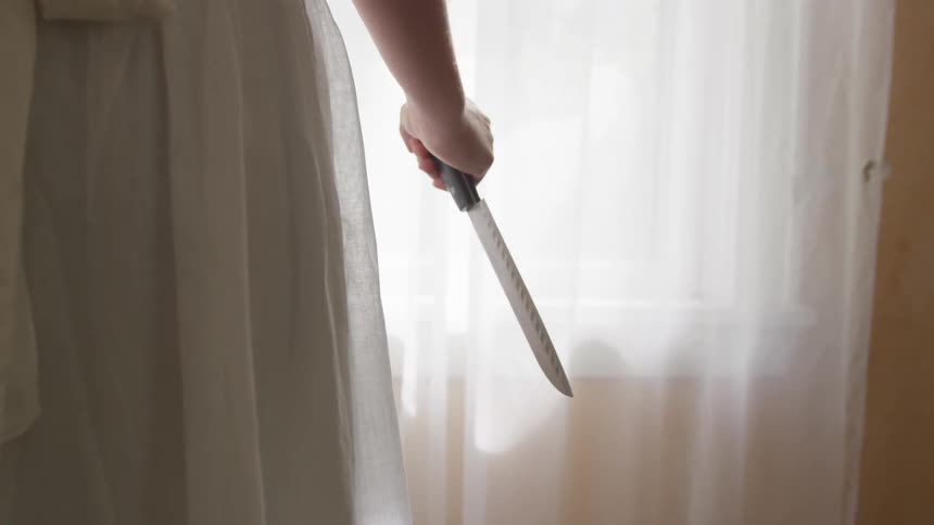 A woman wearing a thin white dress stands in front of a window holding a knife in a stabbing position