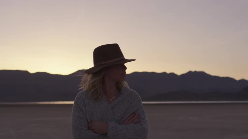 A woman stands in the desert with her arms crossed wearing a hat and a sweater as the sun sets behind the mountain range behind her