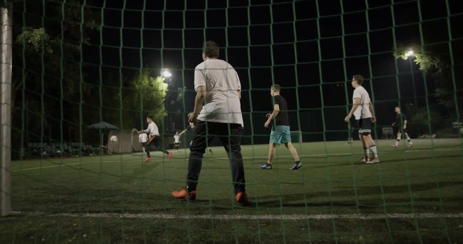 A goalie blocks the ball as men play soccer at night