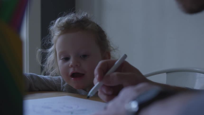 A toddler sitting at a table watching an adult write