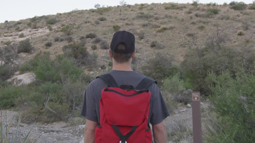 Man wearing cap and  red backpack walks uphill by day