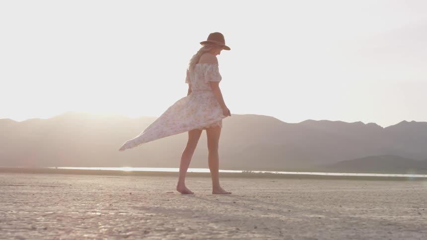 A woman walks over the desert dry cracked soil in her bare feet as her white dress waves in the wind wearing a hat as the sun sets behind the mountains