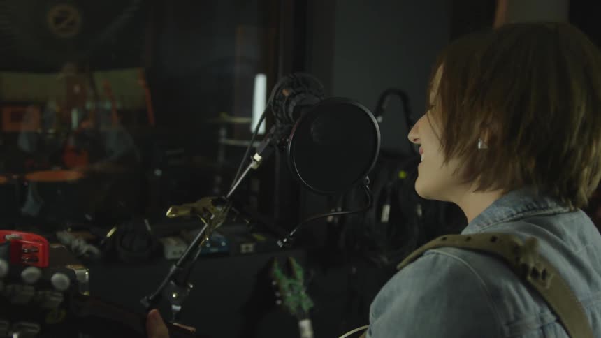 A woman plays guitar and sings into a microphone in a recording studio