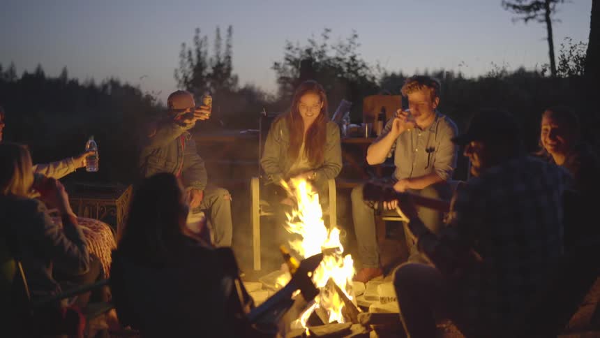A group of young adults are sat around a campfire at dusk raising their glasses to form a toast