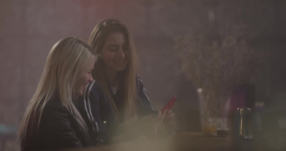 Two young women take selfies sitting at a bar